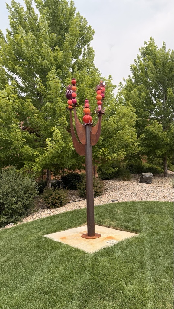 Abundance Tree consists of an 11-foot modern, stylized tree with rusted steel branches and colorful cairn stacks of flowers, leaves and round orbs topping each branch.