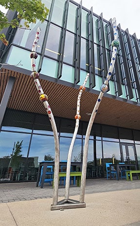 Dancing Sticks by Reven Swanson has three sticks stemming from the base of the sculpture that protrude upwards. Each stick has different colors throughout the upper portion of the stick.